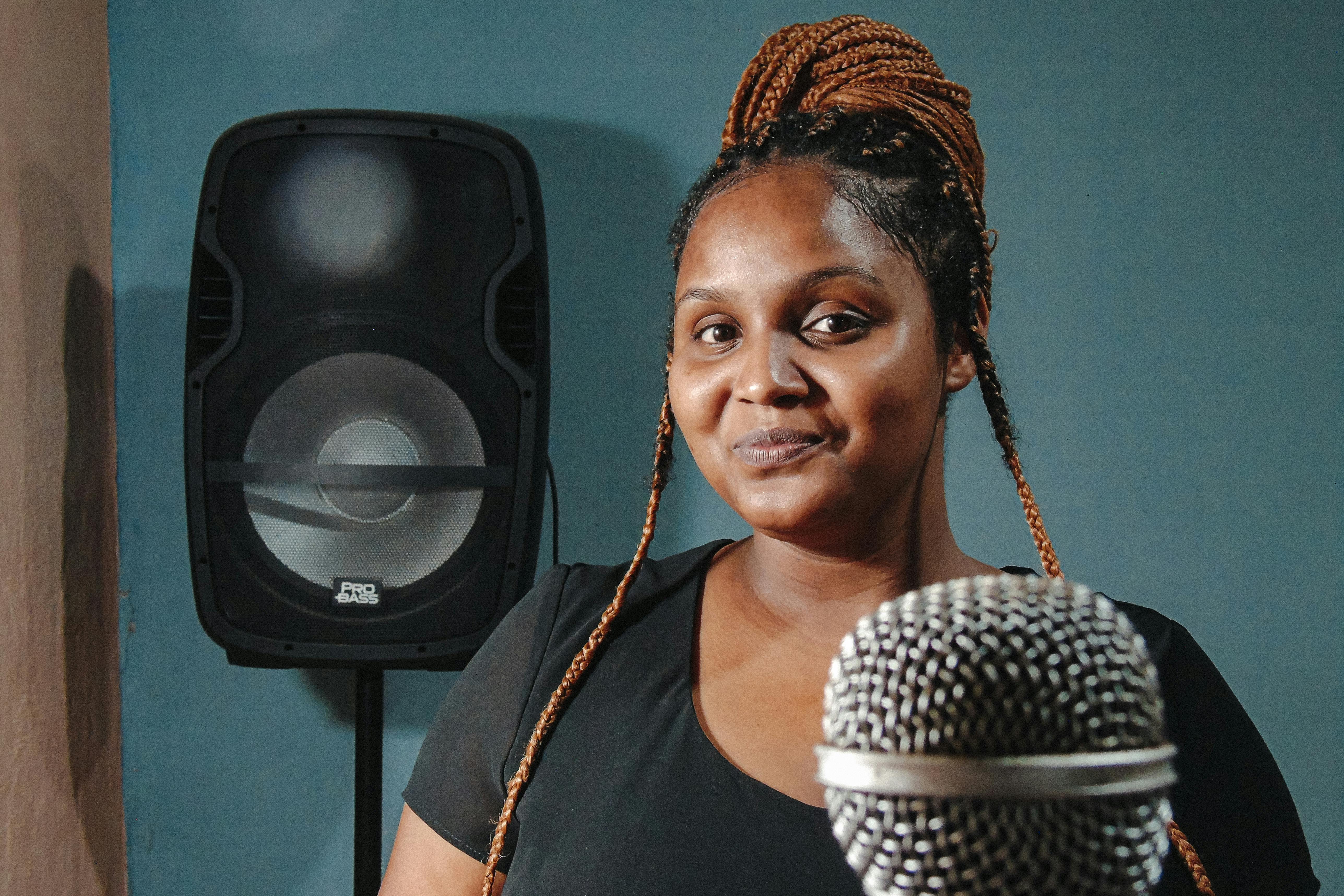 A confident woman with braids smiling in a studio with a microphone and speaker in the background.