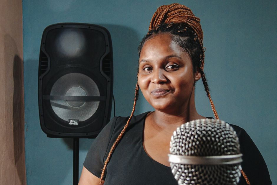 A confident woman with braids smiling in a studio with a microphone and speaker in the background.