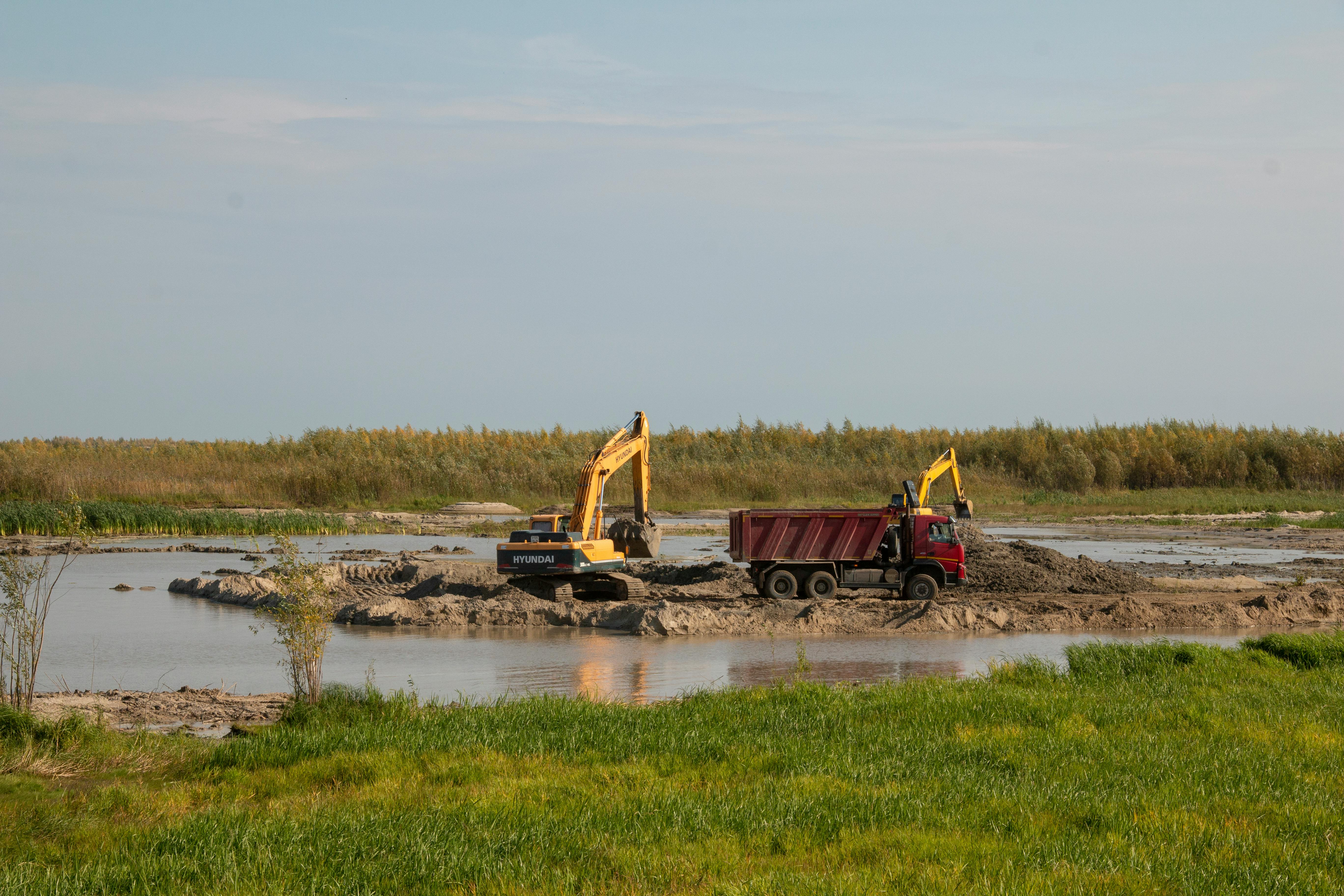 Heavy machinery operating in marshland, excavators and dump truck moving earth by a riverbend.