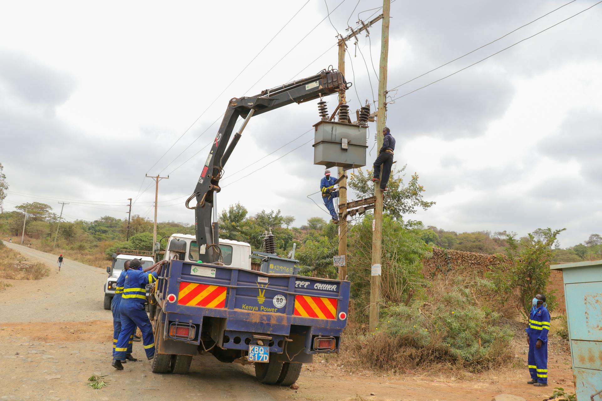 Utility workers in protective gear servicing power lines with a truck crane on a rural road.