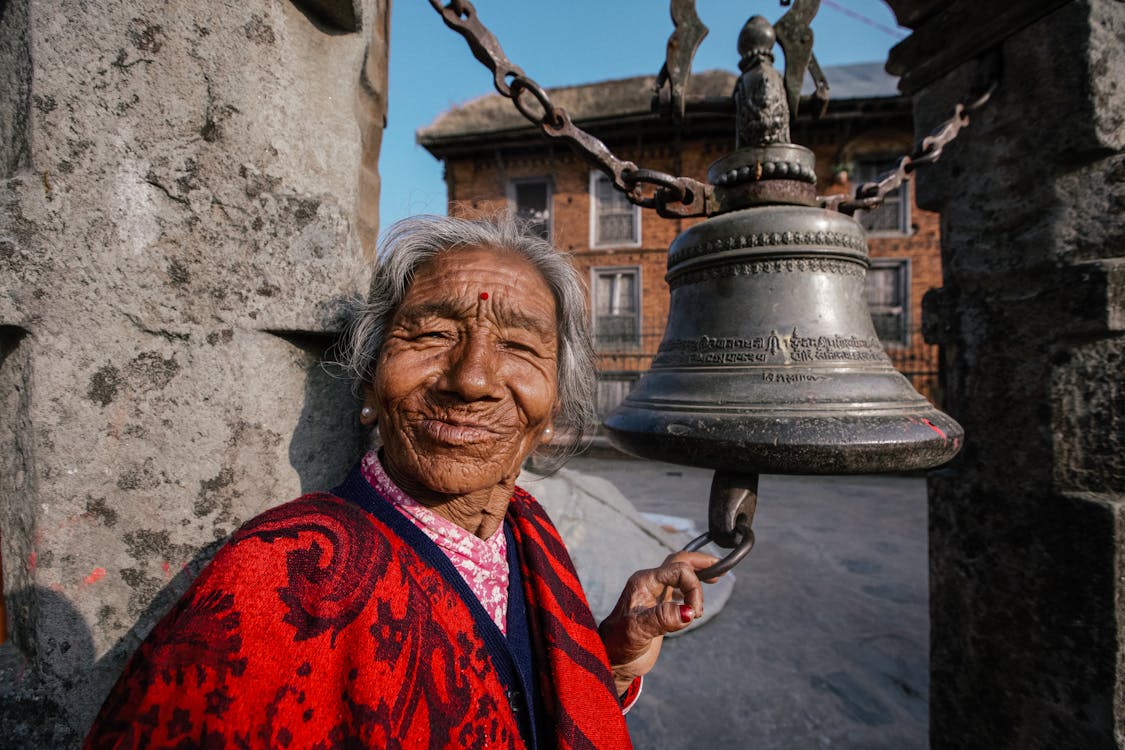 Free Cheerful elderly Hindu female in bright apparel touching aged temple campane and looking at camera on street Stock Photo