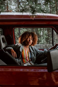 Elegant young woman with afro hair in a red car on a forest road trip, capturing adventure and style.
