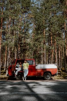 Two people relaxing by a red truck in a dense pine forest on a sunny day.