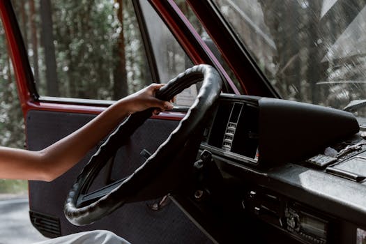 A close-up of a hand gripping a steering wheel in a vintage vehicle, surrounded by forest.
