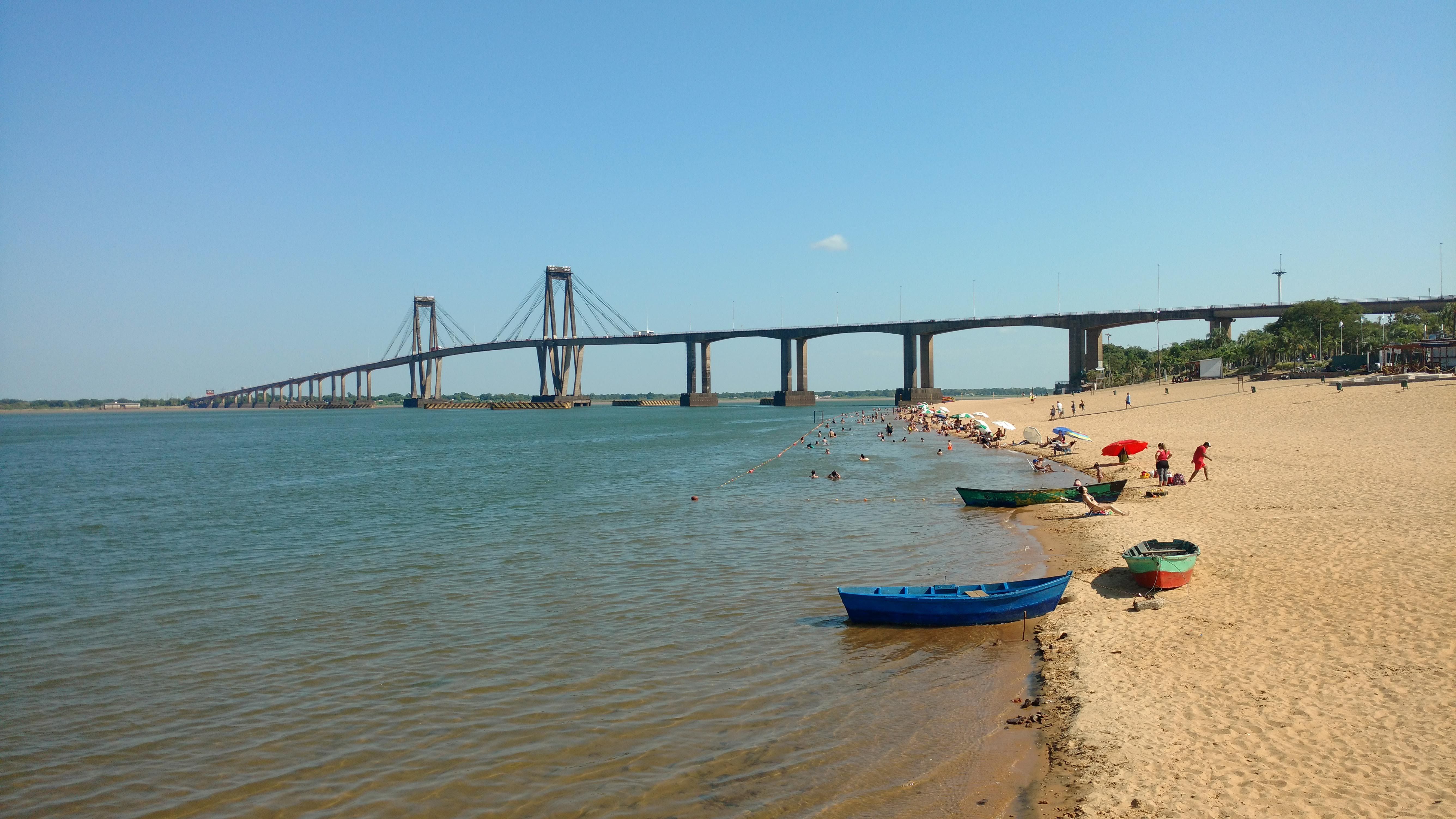 Scenic view of a beach in Corrientes, Argentina with people, boats, and a bridge.