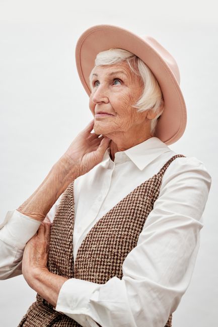 Elegant senior woman with white hair, wearing a pink hat and checked vest, gazing thoughtfully upwards.