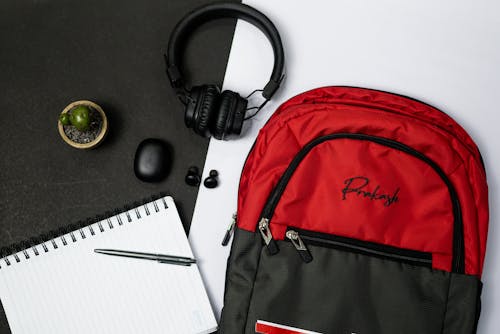 Red and Black Backpack Beside Black Headphones and a Notebook