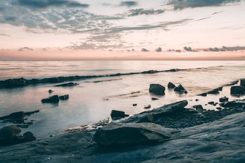 Scenery view of shore with rough stones against rippled ocean with horizon under cloudy sky at sundown