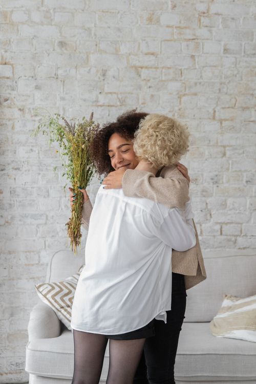 Free African American lady with bouquet of flowers embracing anonymous female friend while standing in room near couch against brick wall Stock Photo
