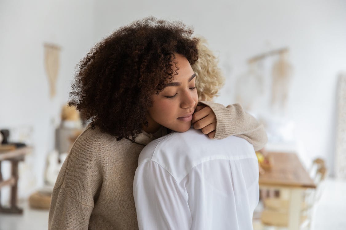 Free African American woman embracing female anonymous friend while standing in cozy light room with wooden table and decorations on wall with blurred background Stock Photo
