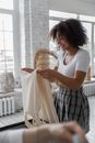 Side view of happy African American female putting dress in suitcase while packing luggage for trip in room with crop person