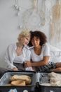 African American lady with closed eyes cuddling female friend while sitting on bed with opened suitcase during packing things in cozy room