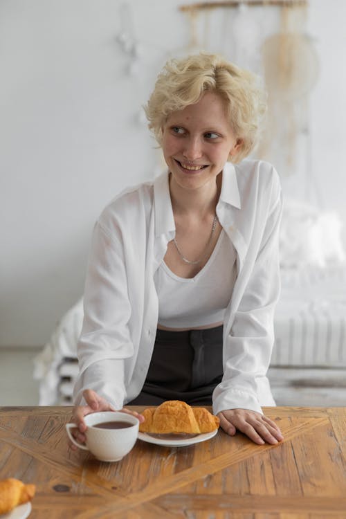 Happy attractive female in trendy outfit sitting at table with fresh croissant and cup of hot coffee while smiling and looking away