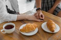Crop unrecognizable girlfriends in trendy clothes holding hands while sitting at wooden table with cup of tea and croissants