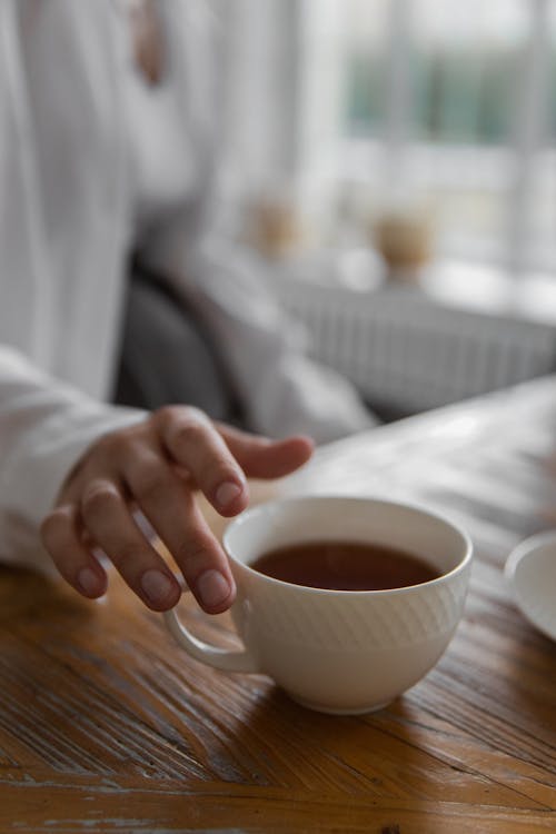 Free Crop faceless person holding cup of hot drink while sitting at wooden table at home Stock Photo
