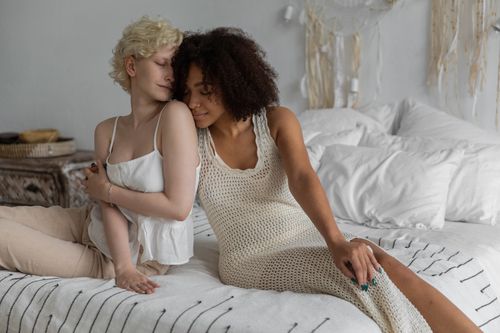 Free Attractive homosexual diverse females in stylish dresses sitting on comfy bed and embracing with eyes closed Stock Photo