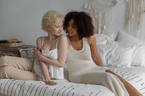 Free Romantic young multiethnic girlfriends in trendy light dresses embracing on comfortable bed and looking at each other with tenderness Stock Photo