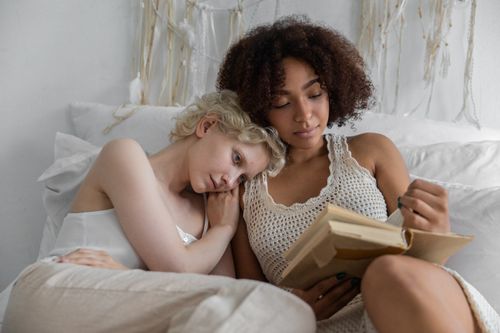 Free Young lesbian diverse couple caressing tenderly while reading book together in cozy bedroom Stock Photo