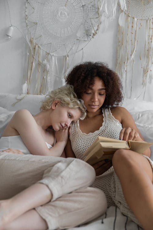 Loving diverse same sex couple relaxing on bed with book