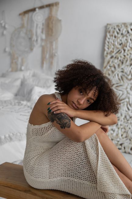 Side view of sad African American female embracing knees while sitting in cozy bedroom