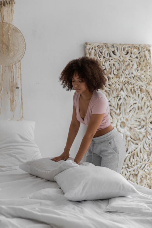 Free Awake African American female making bed in white bedclothes in bedroom in morning Stock Photo