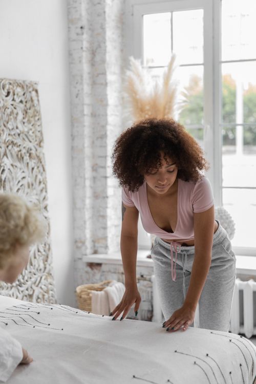 Free African American female adjusting soft blanket while doing housework with lesbian female in cozy bedroom in morning Stock Photo