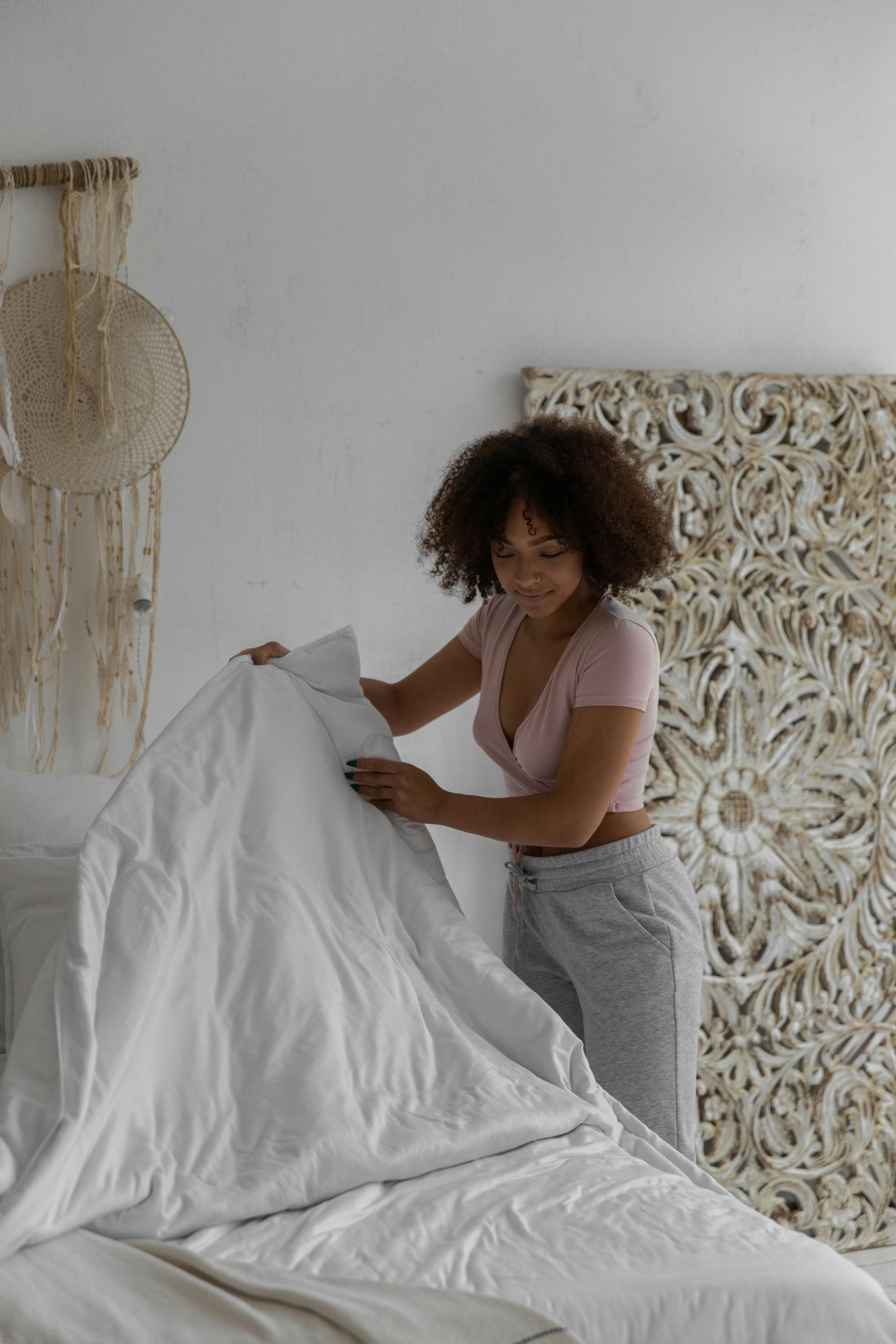 Side view of young fit African American female with curly hair in casual clothes making bed in cozy minimalist room in morning
