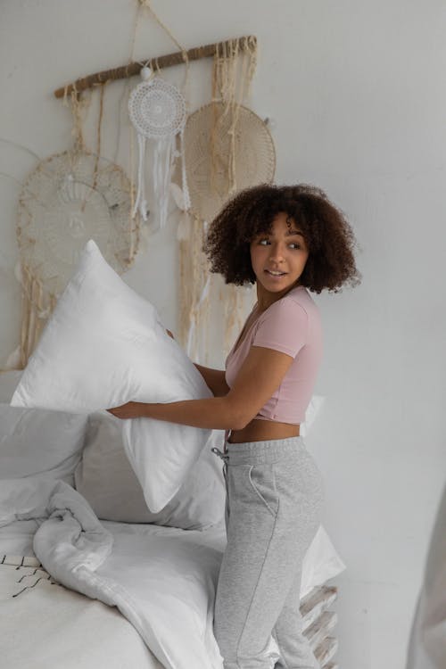 Free Side view of smiling young African American female organizing bed with soft pillows in white bedclothes Stock Photo