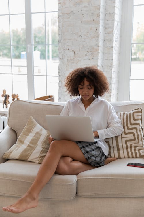 Full body of young stylish barefooted black female freelancer with Afro hair in casual outfit working remotely on laptop while sitting on comfortable sofa in cozy living room