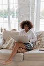 Full body of young stylish barefooted black female freelancer with Afro hair in casual outfit working remotely on laptop while sitting on comfortable sofa in cozy living room