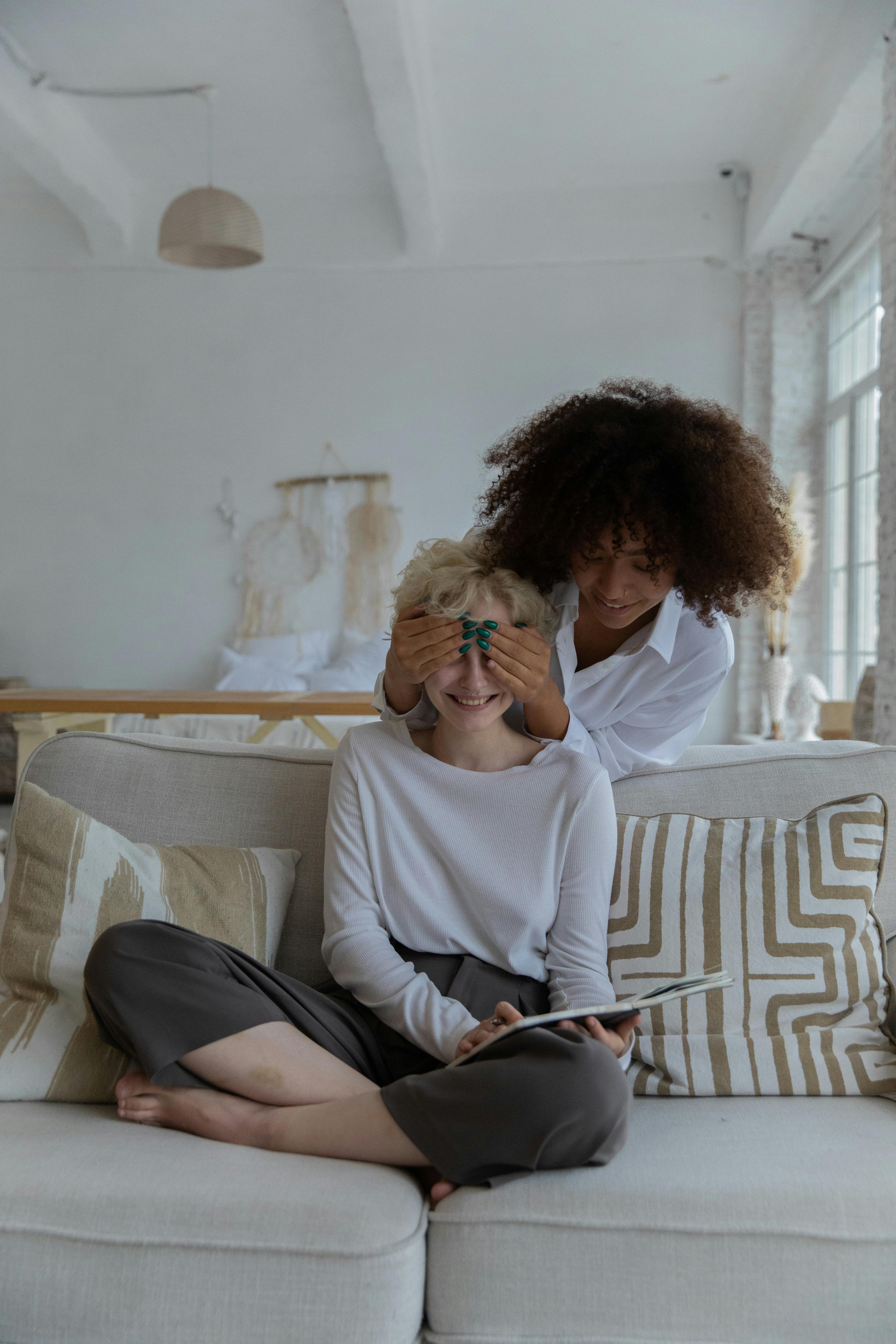 cheerful young black lady covering eyes of girlfriend relaxing on sofa with book