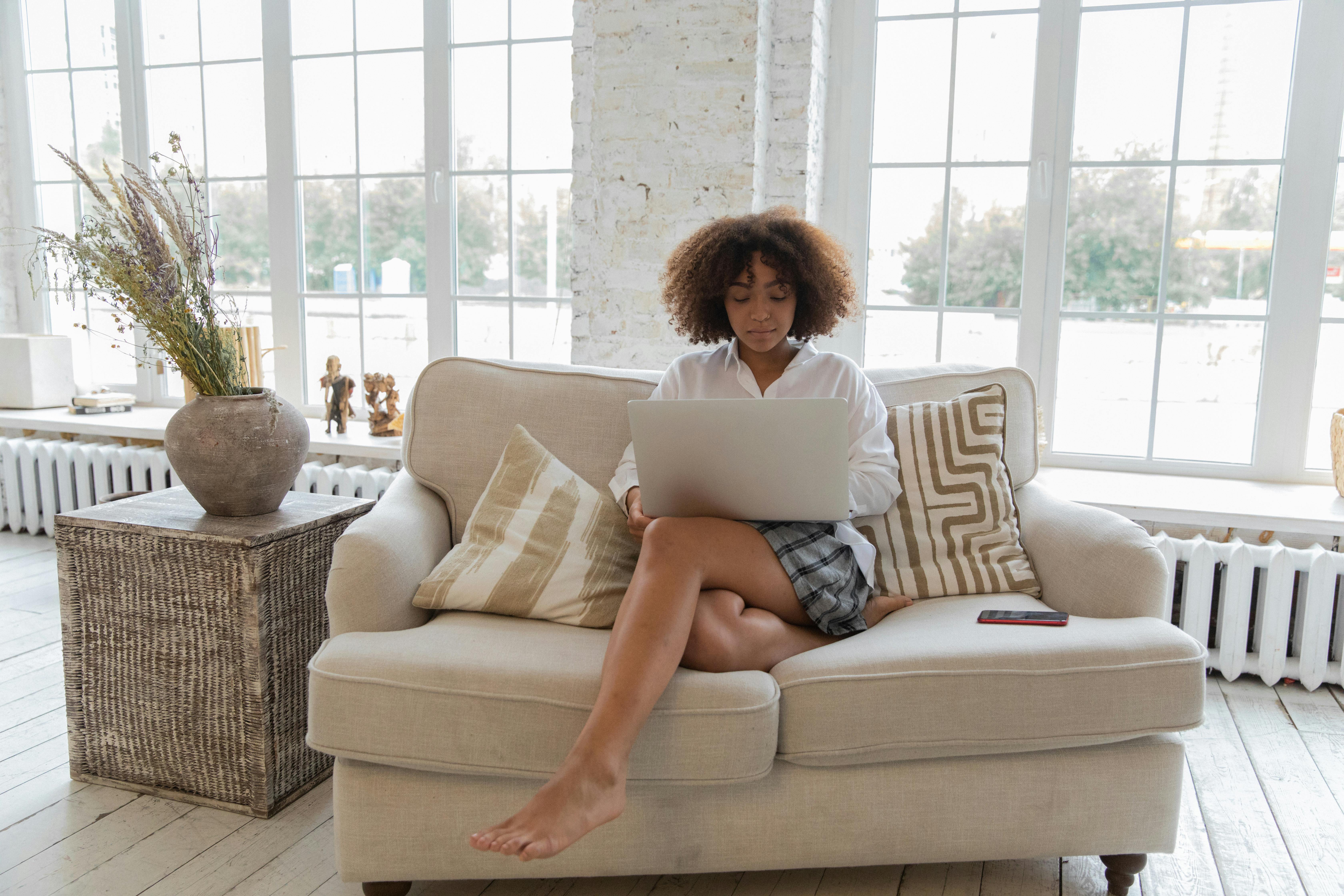 Full length of barefooted  young ethnic female freelancer with Afro hair in casual outfit sitting on comfortable sofa and working online on laptop in stylish apartment