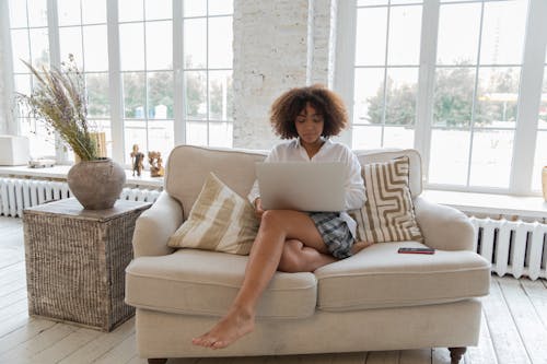 Free Full length of barefooted  young ethnic female freelancer with Afro hair in casual outfit sitting on comfortable sofa and working online on laptop in stylish apartment Stock Photo