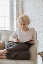 Concentrated young female author with short blond hair in casual outfit taking notes in diary while creating new story at home