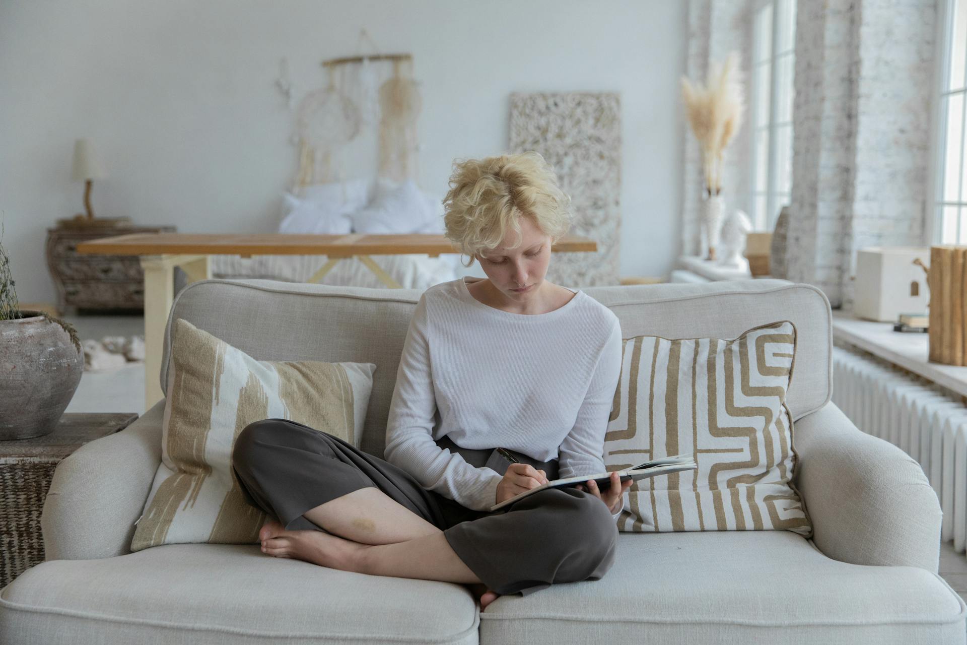 Full body of young barefooted pensive lady with curly blond hair in casual clothes sitting on soft  couch and writing thoughts in notebook during remote work at home