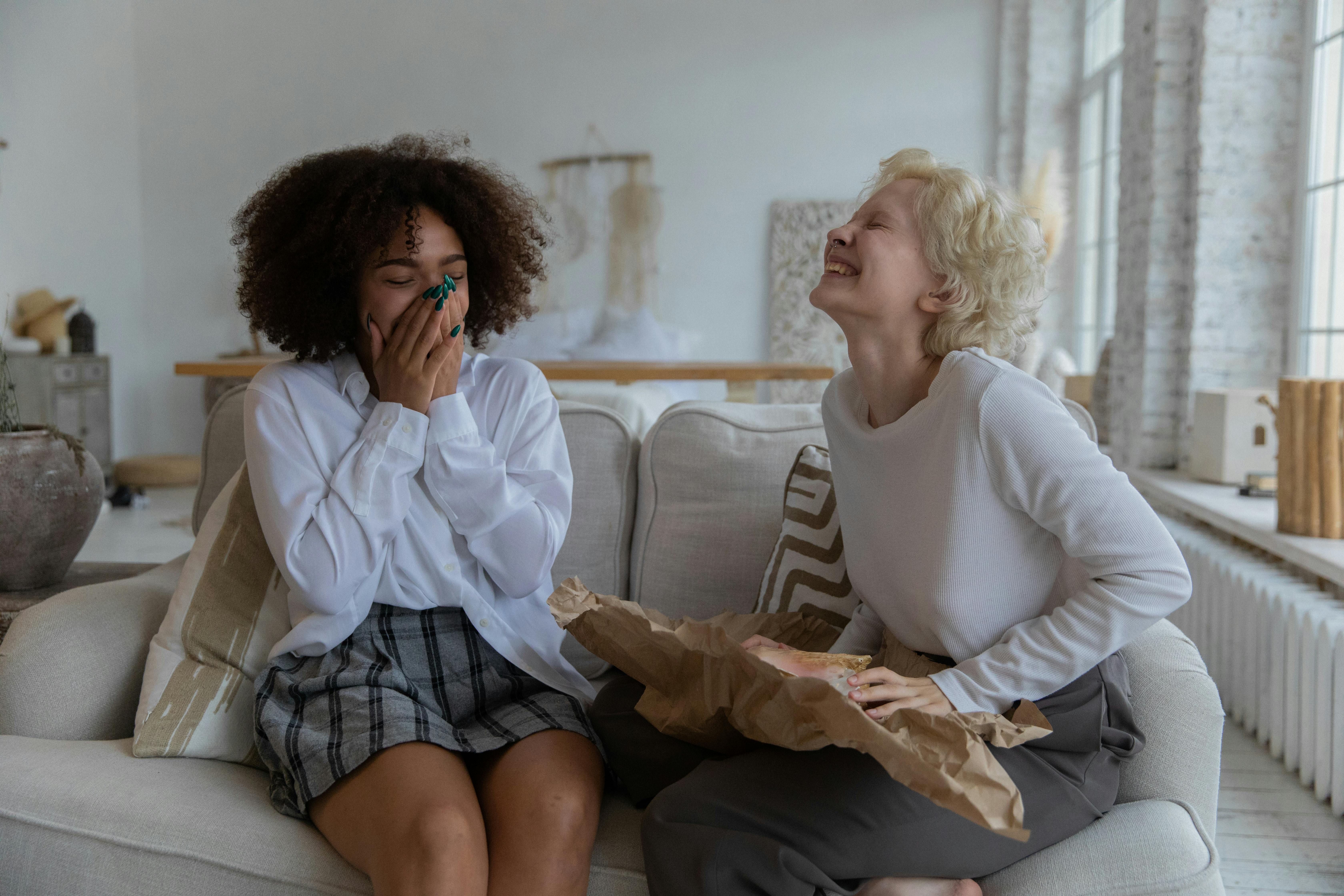 joyful young diverse girlfriends laughing with closed eyes while chilling on couch at home