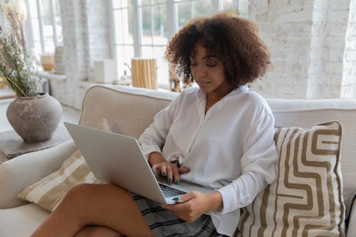 Calm young ethnic lady typing on laptop sitting on couch in cozy apartment