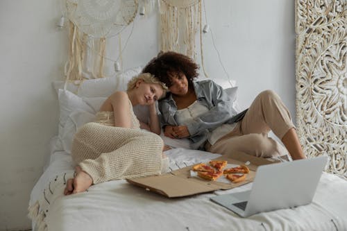High angle of young diverse romantic female homosexual couple lying on soft bed with pizza box and watching interesting movie on laptop during weekend at home