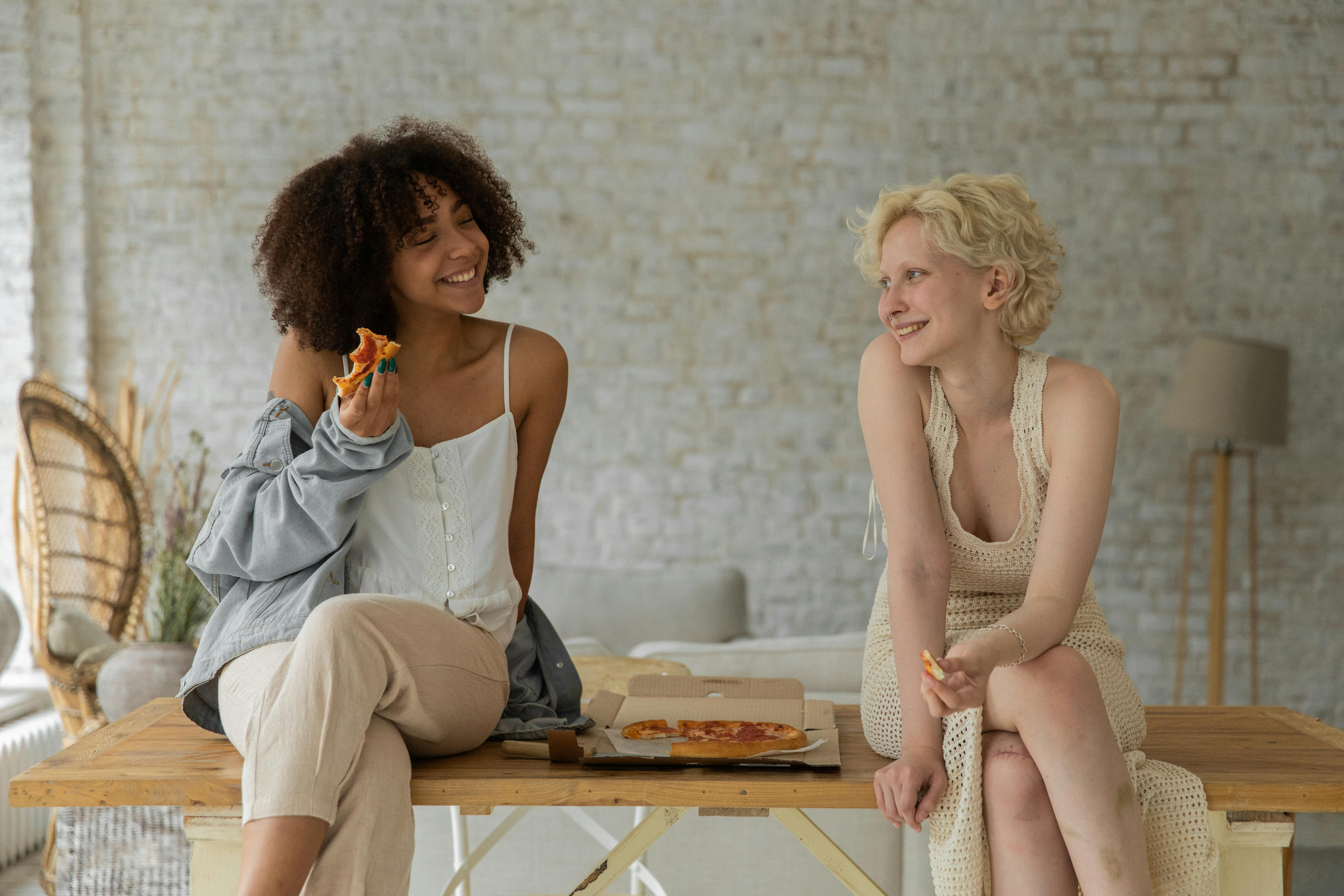 happy young diverse homosexual women eating pizza sitting on table at home