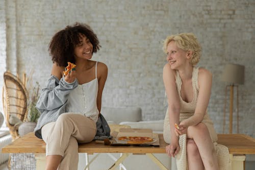 Happy young diverse homosexual women eating pizza sitting on table at home