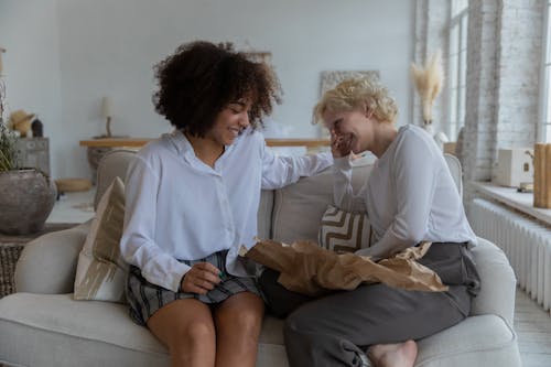 Happy young multiethnic female friends in casual clothes laughing while relaxing on comfortable couch and gossiping together at home