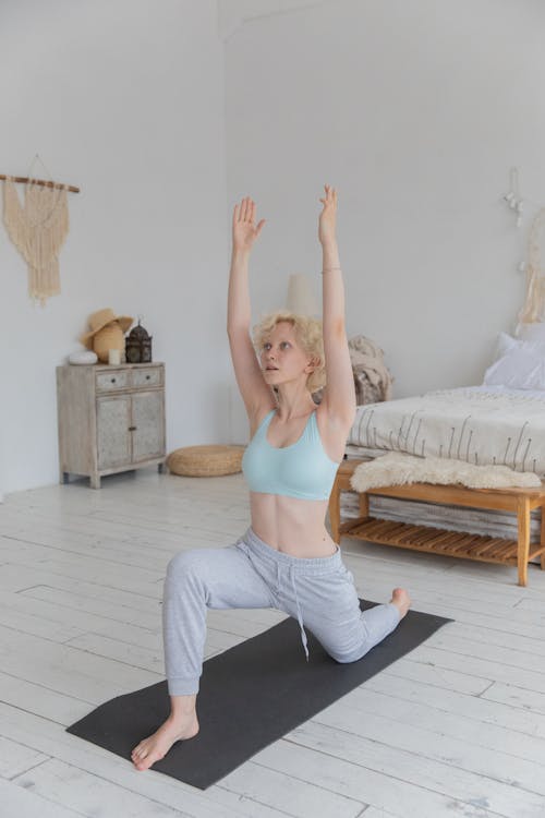 Free Full body of young fit barefooted lady with blond hair in activewear performing Crescent Lunge on the Knee asana while practicing yoga in cozy bedroom Stock Photo