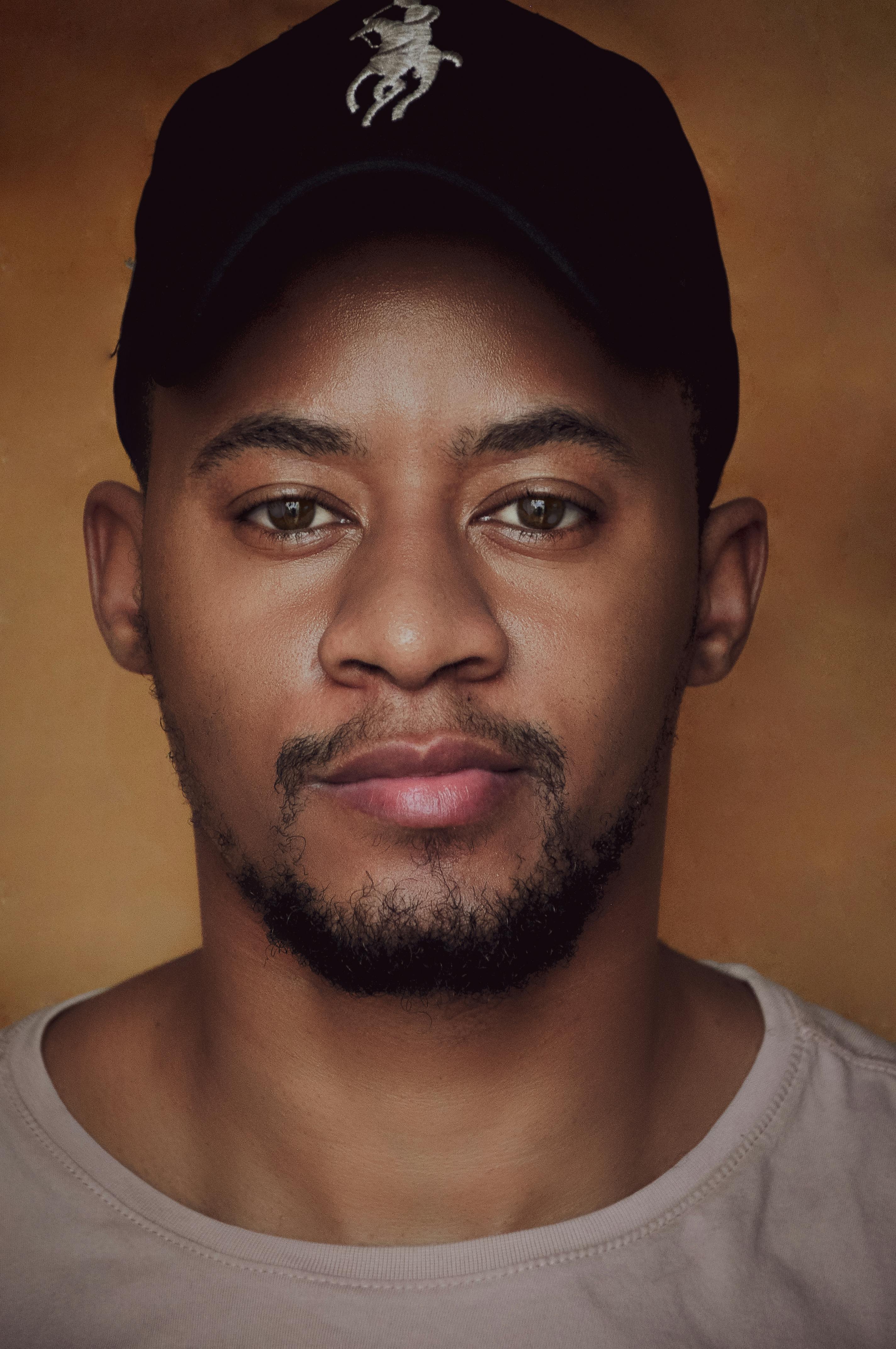 A close-up portrait of a man with a beard wearing a black hat against a warm background.