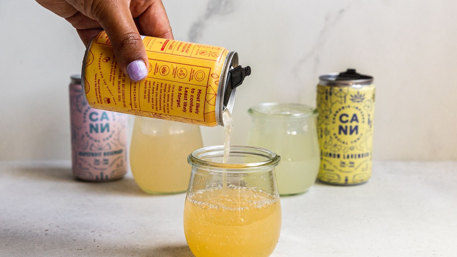 Hand pouring cannabis-infused beverage from a can into a glass, set against a marble backdrop.