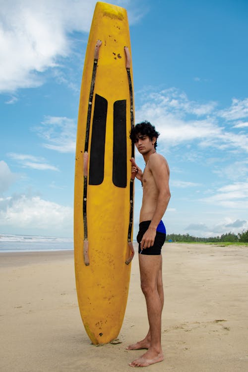 Free Full body of male in swimsuit looking at camera while standing on sandy beach with surfboard near sea against cloudy sky in tropical resort Stock Photo