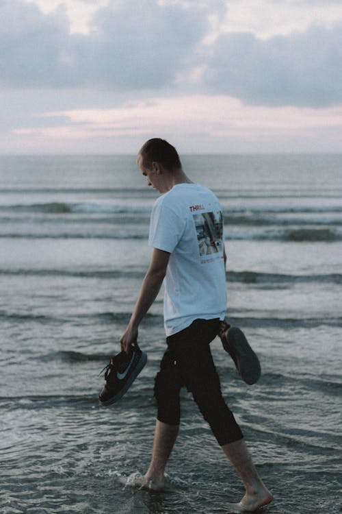 Back view of anonymous male tourist carrying sneakers while walking on wavy ocean under cloudy sky at sundown