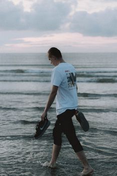 Back view of anonymous male tourist carrying sneakers while walking on wavy ocean under cloudy sky at sundown
