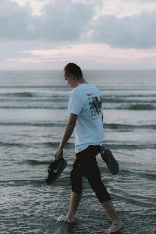 Free Back view of anonymous male tourist carrying sneakers while walking on wavy ocean under cloudy sky at sundown Stock Photo