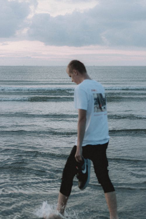 Back view of anonymous barefoot tourist strolling on rippled sea under cloudy sky in twilight