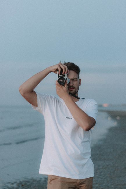 Serious bearded male in casual wear taking photo and standing on coast near waving sea under cloudy sky in gloomy weather